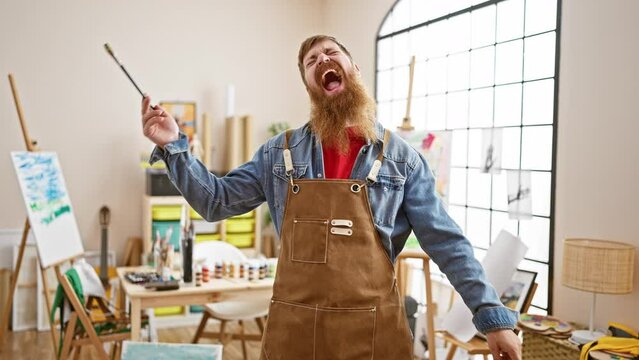 Upbeat, young redhead artist guy, joyfully swinging his paintbrush to the music in art studio, revealing his playful side, crafting with paint, under the influence of his irish heritage.
