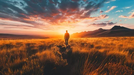 Foto op Aluminium  a man standing on top of a dry grass covered field under a cloudy blue and pink sky with the sun setting in the distance behind him and a mountain range in the distance. © Jevjenijs