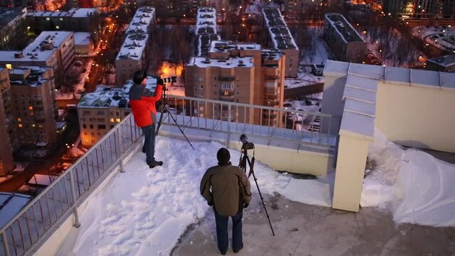 Two photographers shoots the evening city from the roof using a tripod