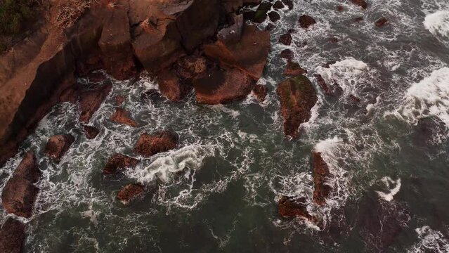 Top static view of texture waves of ocean crashing against stones coastline in the warm sunset light. The sea surf breaking on rocks and turning into a seething white foam. Amazing calm aerial footage