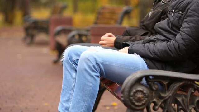The legs and hands of two men sitting on a bench in autumn park