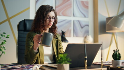 Business woman holding coffee sitting office desk close up. Girl looking laptop.