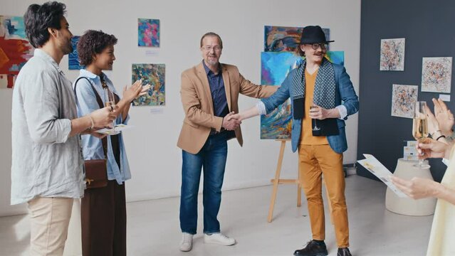 Medium full shot of reception during exhibition opening at art gallery - owner greeting eccentric male artist, shaking hands, cheerful artist welcoming guests and demonstrating new painting on easel