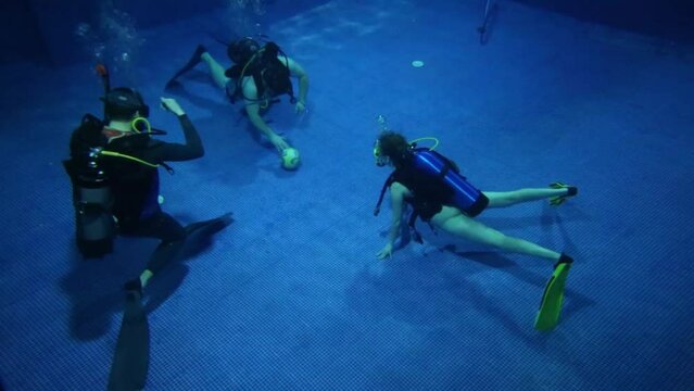 Three divers with scubas play with ball underwater on bottom of pool