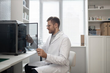 Smiling Caucasian male technician looking at 3D bone model in hand while opening enclosed 3D...