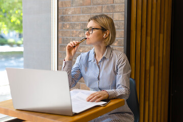 A woman in a study at a desk with a laptop and papers looks thoughtfully out the window. Business in glasses lady working in office, accountant, woman in glasses looking thoughtfully out the window