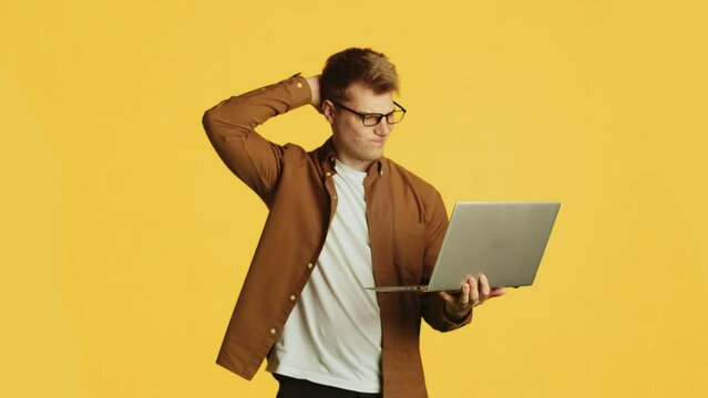 Young handsome caucasian man in glasses and casual clothes standing with a laptop, scratching the back of his head and looking perplexed on a yellow background in the studio