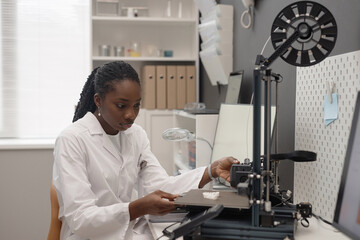 Medium shot of Black woman technician examining produced sample on 3D printer hot bed while sitting in modern laboratory