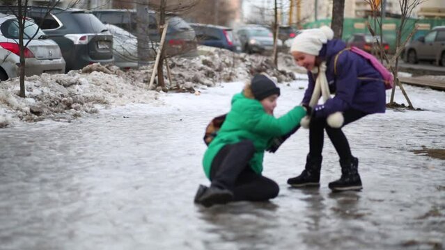 Boy runs, slips and falls down on icy surface, girl helps him