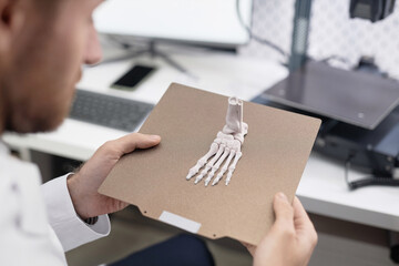 Over the shoulder shot of printed 3D human foot bone prototype on top of aluminum print bed in hands of male laboratory technician