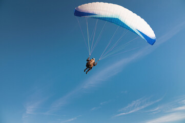 A paraglider takes off from a mountainside with a blue and white canopy and the sun behind. A paraglider is a silhouette. The glider is sharp, with little wing movement.