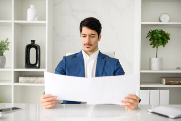 A focused Hispanic businessman or male office employee in a blue suit scrutinizes documents,...
