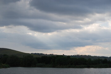 Wild steppe nature. Trees, bushes, grass with green foliage grow on a hillside under a sky with gray clouds. A small pond with light gray water at the bottom. The sun's rays break through the clouds.