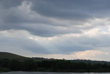 Wild steppe nature. Trees, bushes, grass with green foliage grow on a hillside under a sky with gray clouds. A small pond with light gray water at the bottom. The sun's rays break through the clouds.