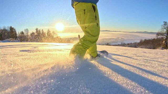 POV: Unrecognizable snowboarder guy riding powder snow, dog running next to him at golden winter sunrise. Freeride snowboarder carving fresh snow in backcountry with happy dog running along at sunset