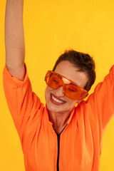 Middle age funny and Cheerful woman wear modern Orange overalls with glasses standing over yellow color background. Female having fun and energetic movement studio shot 