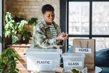 Young university student is managing waste sorting at home, smiling. Recyclable materials as paper,...