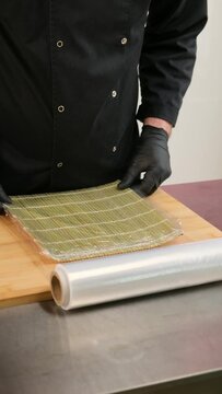 Vertical video: Close-up of a chef's hands wrapping a bamboo mat in cling film to prepare sushi rolls.