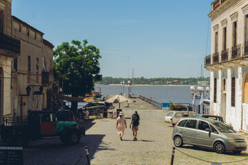 woman and man walk through a coastal city with a pier and the sea in the background, Colonia del...