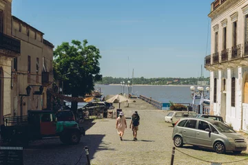 Zelfklevend Fotobehang woman and man walk through a coastal city with a pier and the sea in the background, Colonia del Sacramento © juanpablo