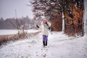 Fototapeta na wymiar a small child in winter clothing joyfully running on a snowy path lined with bare trees and autumn leaves