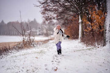 Fototapeta na wymiar a small child in winter clothing joyfully running on a snowy path lined with bare trees and autumn leaves