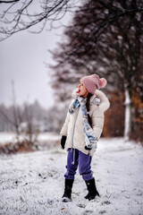 young girl standing in a snowy landscape, looking to the side with a backdrop of bare trees and falling snowflakes.