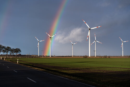 Wind turbines against dark cloudy sky after a thunderstorm. A big rainbow in the middle of the wind farm. Wind turbines are brightly lit. Country road along fields.