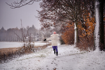Fototapeta na wymiar The photo captures a child in a white coat and purple pants joyfully running in a snowy landscape, with snowflakes gently falling around.