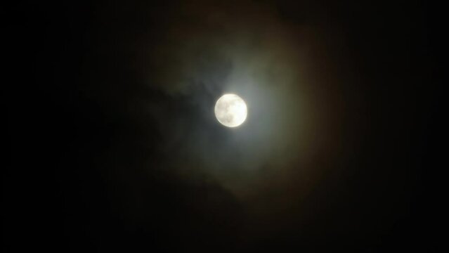 Full moon in the dark night sky with fast flying clouds. The lunar corona is a light optical phenomenon in the form of colored clouds around the Moon