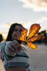 Hispanic Young Woman Holding a Yellow Leaf in Focus with Sunset in the Background. An out-of-focus...