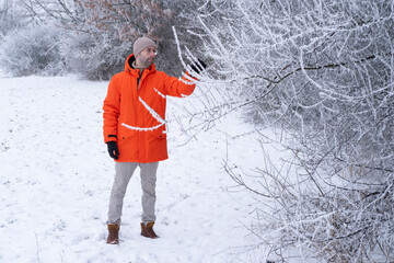thoughtful charismatic man 30 years old in bright orange jacket, in glasses, fir trees in snow,...