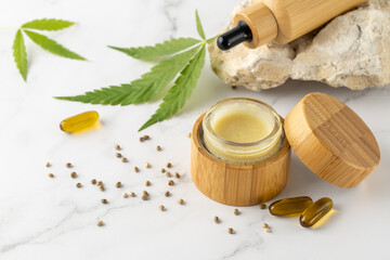 Obraz na płótnie Canvas Hemp salve for joints and muscles in open glass wooden jar and relaxing CBD serum oil in dropper bottle and in capsules as a complex in the treatment of the musculoskeletal system