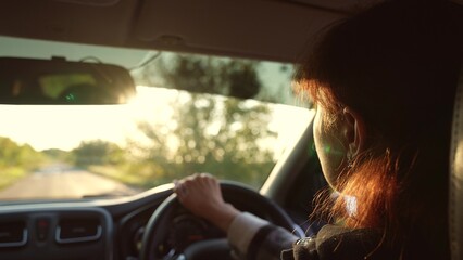 Fototapeta na wymiar Redhead woman driver driving car at sunlight countryside rural summer landscape road trip closeup back view. Modern female riding automobile holding steering wheel enjoy automobile movement on path