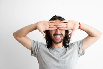 young adult Latin man with dental braces smiling while covering his eyes with his hands, studio...