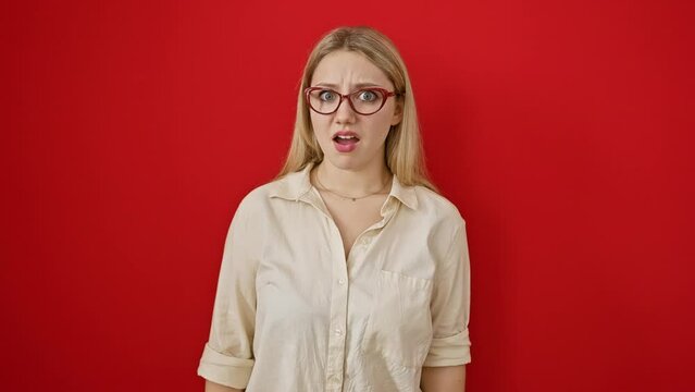 Young blonde woman, skeptical and sarcastic, standing in disbelief over a red isolated background, with a cynical look and open mouth expression of shock