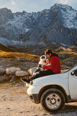 A man sitting on the hood of his off-road car with his border collie dog with the mountains in the background. Adventure travel. traveling with pet. © Alberto