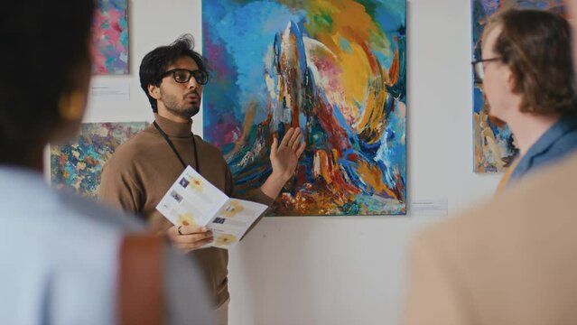 Medium shot of confident handsome young Arab male art museum guide talking to unrecognizable visitors at contemporary art exhibition telling them history and idea of abstract painting