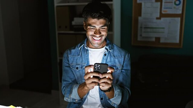 Handsome young hispanic man smiling at phone screen in office at night