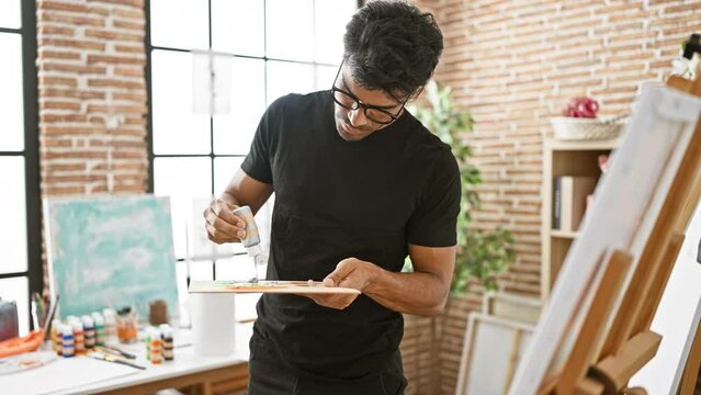A focused man with glasses squeezing paint onto a palette in a bright art studio.