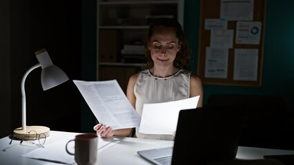 A focused woman working late in a dimly lit office, reviewing documents by a laptop and desk lamp.