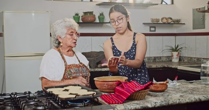 Latina grandmother and granddaughter chat while grandmother teaches her granddaughter how to cook tortillas.