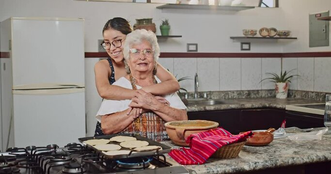 Teenage granddaughter hugs her grandmother and laughs in front of the camera, in the kitchen of their home.