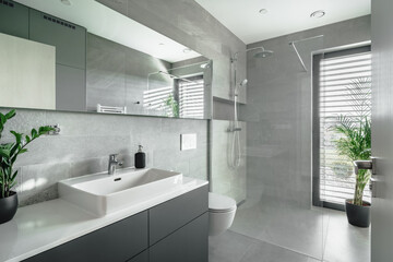 Fototapeta na wymiar The image shows a modern bathroom with grey tiles, a white sink, mirror, toilet, and a glass shower. There are two green plants for a natural touch. Natural light comes in through a window.