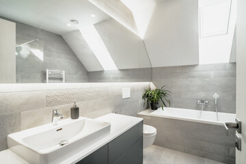The image shows a modern, well-lit bathroom with grey tiles. It has a sink, mirror, toilet, and...