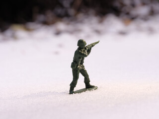 Fototapeta na wymiar War toy soldier in bright daylight on a cold, snowy battlefield landscape. Frozen earth mounds, trees in the blurred dark background.