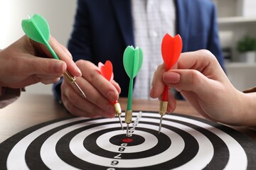 Business targeting concept. People with darts aiming at dartboard at table indoors, closeup
