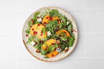 Tasty salad with persimmon, blue cheese, pomegranate and walnuts served on white tiled table, top...