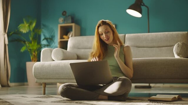 Excited beautiful woman having video call on laptop while sitting on the floor leaning back on couch in living room