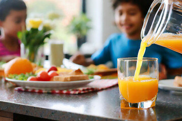 Selective focus on orange juice pouring from jug into glass with little children in background